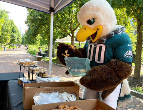 Philadelphia Eagles Mascot Swoop holding Green2Go Container at table full of soft pretzels